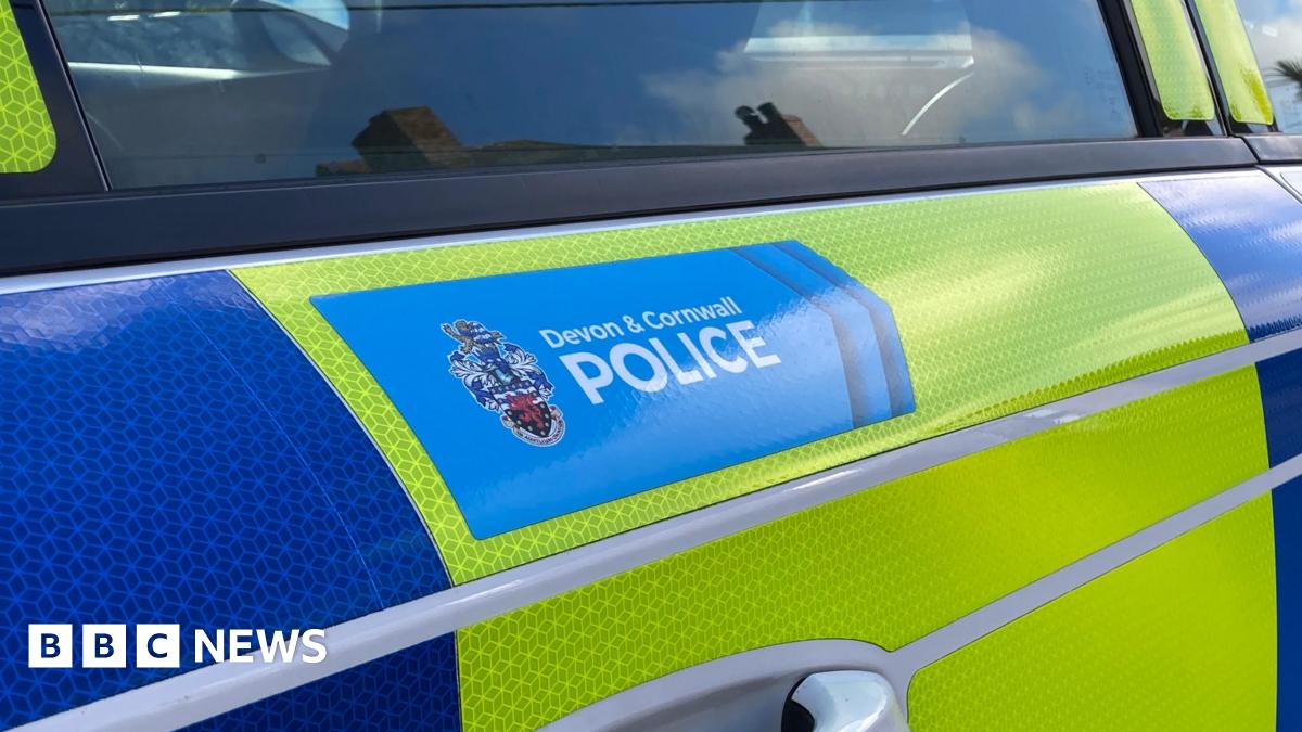 The side of a yellow and blue police vehicle. There is a blue section with the words 'Devon and Cornwall Police' inside of it. The door handle is below and a window above the section.