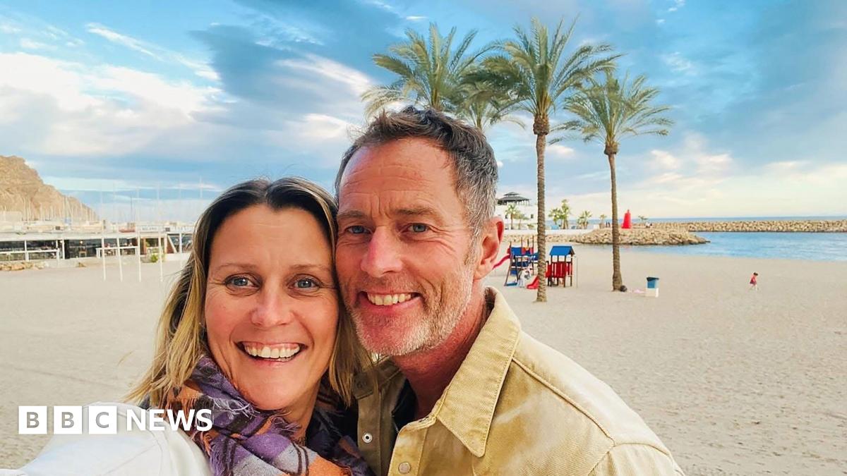 A couple on a beach. It is sunny and the sand is white. Behind them are some trees.