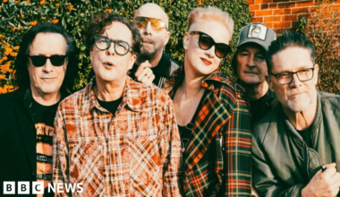 The band pose outside with a wall and greenery behind them. Three have sunglasses on and two are wearing checked shirts with another in a black jacket and a fourth member in a green jacket. Two others are obscured in the photo.