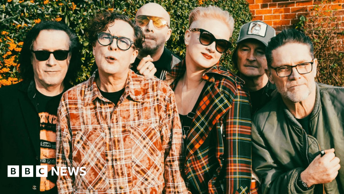 The band pose outside with a wall and greenery behind them. Three have sunglasses on and two are wearing checked shirts with another in a black jacket and a fourth member in a green jacket. Two others are obscured in the photo.