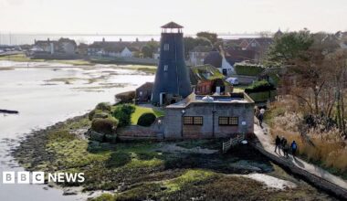 A wide, aerial shot of a part of King Charles III England Coast Path in Hampshire shows four people walking on a path by sea shoreline on a sunny day. The stretch of land between the sea and the path looks muddy and is covered in green moss and low grass. On the other side of the path, there is brown grassland with a few trees with a green field in the background. In the middle of the photo, there is a stone building and a round tower.
