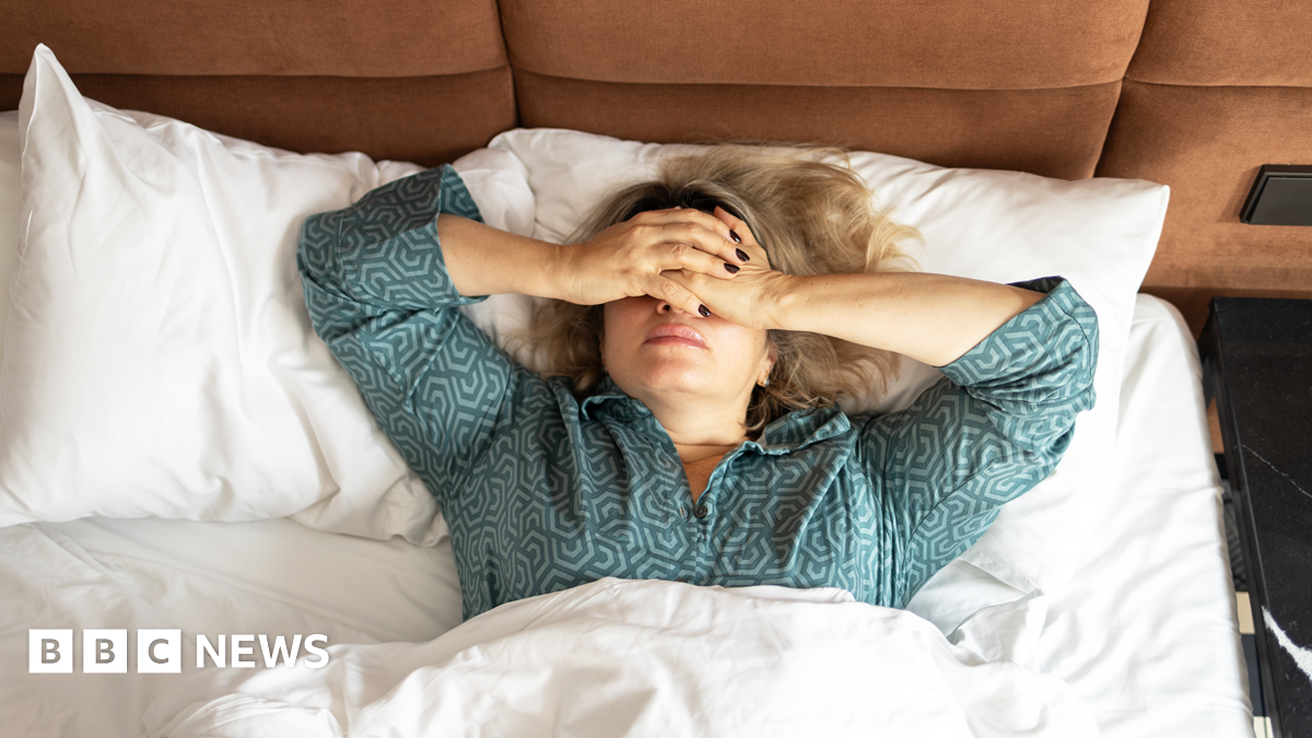 A women in green pajamas lies in bed with white bed linen photographed from above. She has her hands over her eyes.
