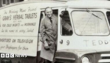 John Gray stands in the door of an old-fashioned van with The working man's friend Gray's Herbal Tablets for Cold Nights and Mornings written on the side. Gray is smiling and wearing a long apron, shirt and tie.