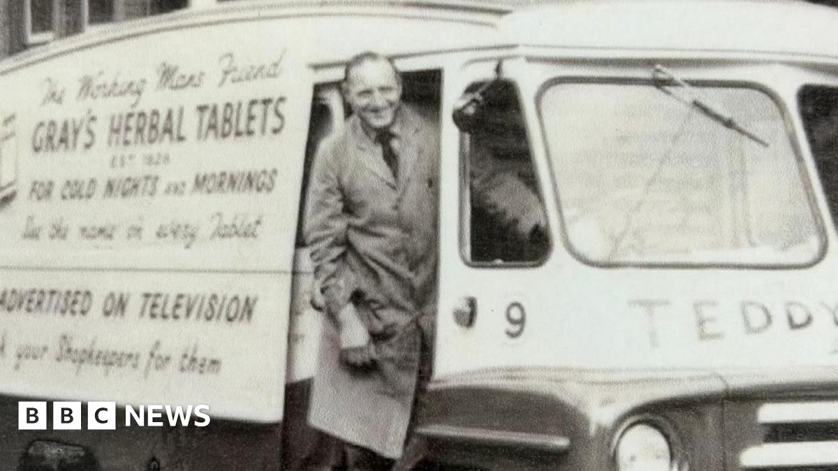 John Gray stands in the door of an old-fashioned van with The working man's friend Gray's Herbal Tablets for Cold Nights and Mornings written on the side. Gray is smiling and wearing a long apron, shirt and tie.