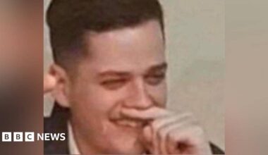 A man holds his finger under his nose while holding his right arm in the air