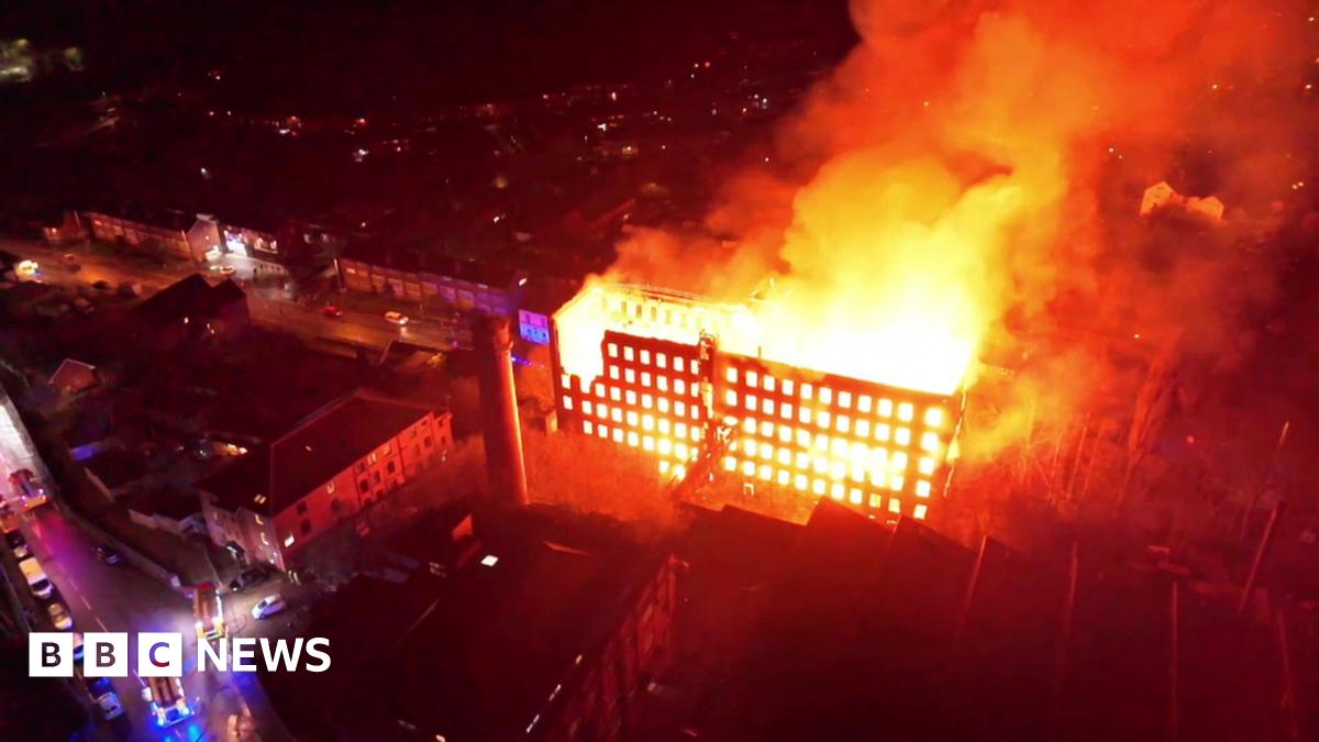 Three residents look on as fire crews tackle a fire in a building at night. One is wearing a bright yellow woolly hat. Behind police tape, a ladder platform is raised up to the third storey of a six-storey building, with jets of water coming from a house.