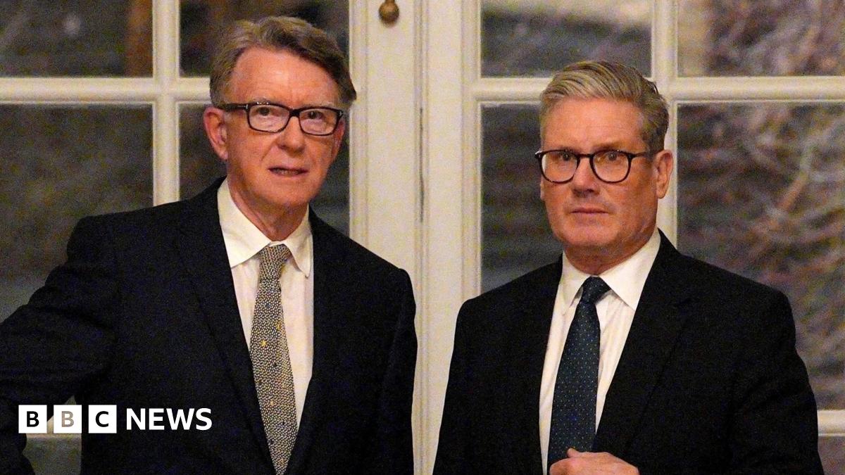 Peter Mandelson stands with Sir Keir Starmer in front of a set of double doors with white panes. Both men wear black suit jackets, white shirts and dark ties.