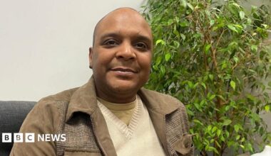 Maurice Blake, who is black, is sitting on a grey couch in front of a plant. He is smiling at the camera.
