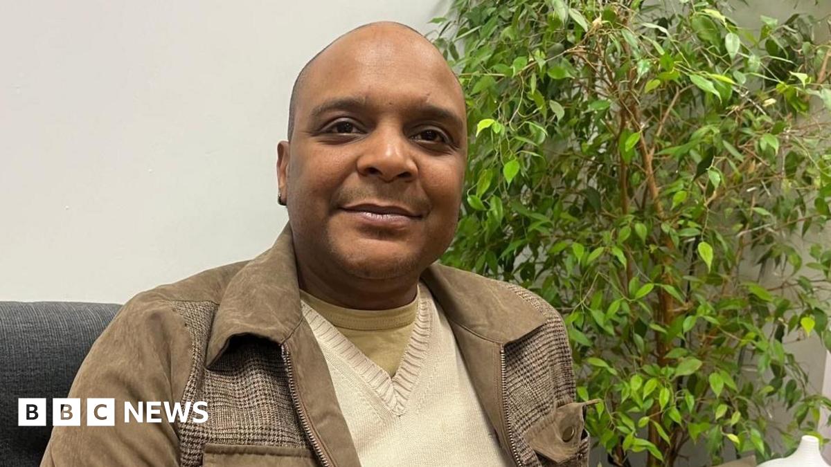 Maurice Blake, who is black, is sitting on a grey couch in front of a plant. He is smiling at the camera.