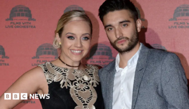 The Wanted singer Tom Parker, wearing a white shirt and grey suit with no tie, with his arm around his smiling blonde wife Kelsey. who is wearing a black and gold dress