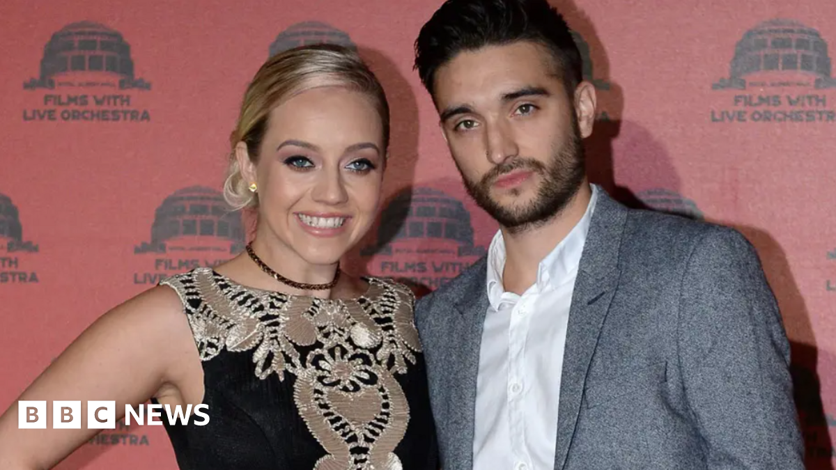 The Wanted singer Tom Parker, wearing a white shirt and grey suit with no tie, with his arm around his smiling blonde wife Kelsey. who is wearing a black and gold dress