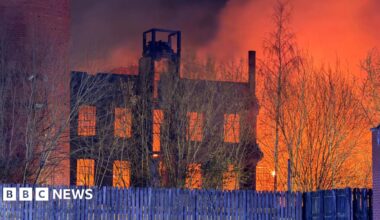 A large blaze at night in a mill building. The sky is lit up orange. Wooden fencing is in front of the building.