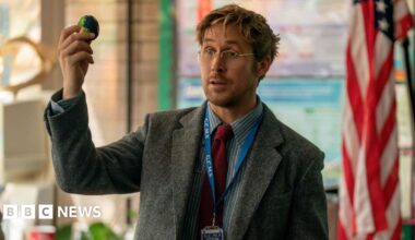 Picture of Ryan Gosling in a still from Project Hail Mary. He is wearing a suit jacket, shirt, tie and glasses and is holding a small globe toy.