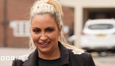 Jodie Marsh has blonde hair in a high pony tail. She is wearing a black top and a black blazer with silver detail on the shoulders. Behind her is a building and a parked white car.