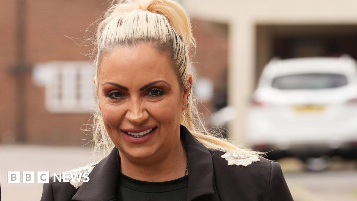Jodie Marsh has blonde hair in a high pony tail. She is wearing a black top and a black blazer with silver detail on the shoulders. Behind her is a building and a parked white car.