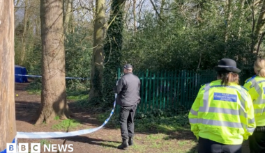 Two female police officers wearing yellow high-vis jackets watch a male officer, wearing black, stand by police tape. In the distance a blue police tent can be seen behind trees in the park