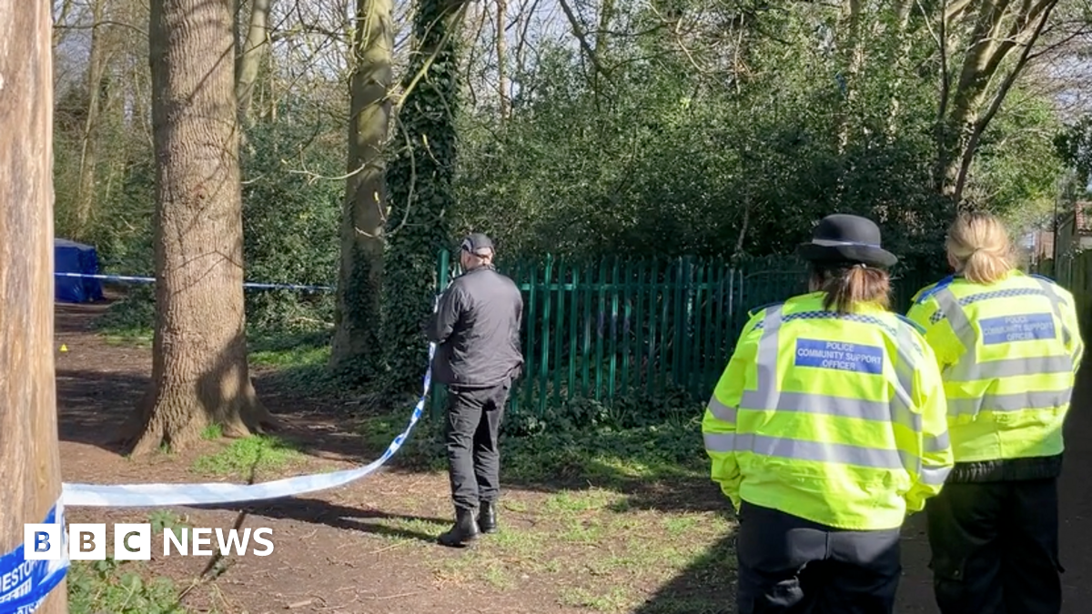 Two female police officers wearing yellow high-vis jackets watch a male officer, wearing black, stand by police tape. In the distance a blue police tent can be seen behind trees in the park