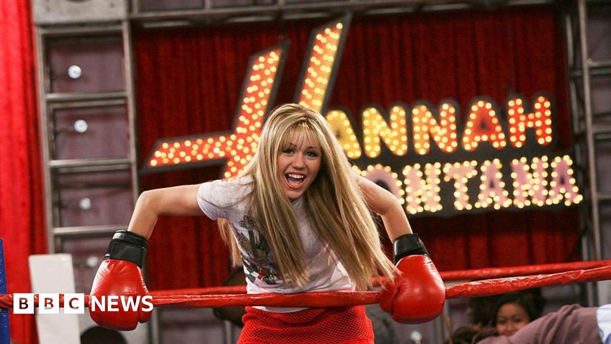 Miley is wearing a long blond haired wig. She is in a red boxing ring holding the edge clad in red boxing gloves. Big letters with lights behind her read Hannah Montana.
