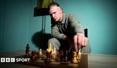 Erling Haaland looks into the camera as he moves a white bishop on a chess board.