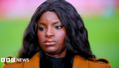 Eniola Aluko looking off-camera. She has long black hair and is wearing an orange jacket.