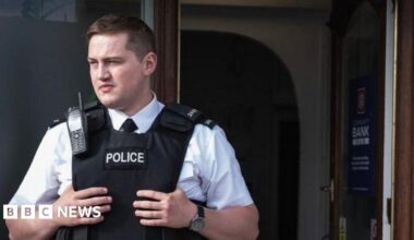 Finnian Garbutt, a man with dark hair, wearing a white shirt with black tie and a black support vest with the words police on it.  He is wearing a black watch on his left wrist and there is a brown wooden door in the background.