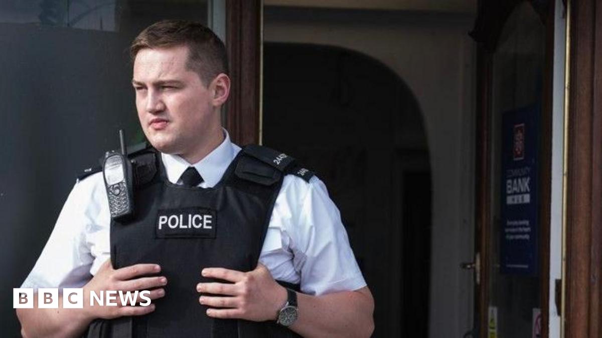 Finnian Garbutt, a man with dark hair, wearing a white shirt with black tie and a black support vest with the words police on it.  He is wearing a black watch on his left wrist and there is a brown wooden door in the background.