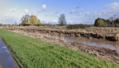 A river running next to a rural single-carriageway road. A short grass verge separates the road from the water.