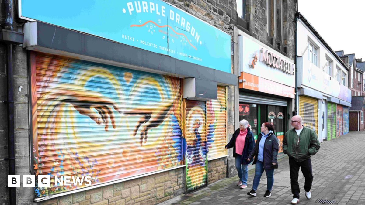Gift Shop Purple Dragon's shutters are down. They are painted in yellow, orange and blue and who two hands reaching out to one another. Two women in jeans and dark coats are walking past and looking at the artwork. Lee Ferry is walking next to them. His hands are in the pockets of his khaki bomber jacket. He has short white hair, a beard, black circular glasses and black jeans.