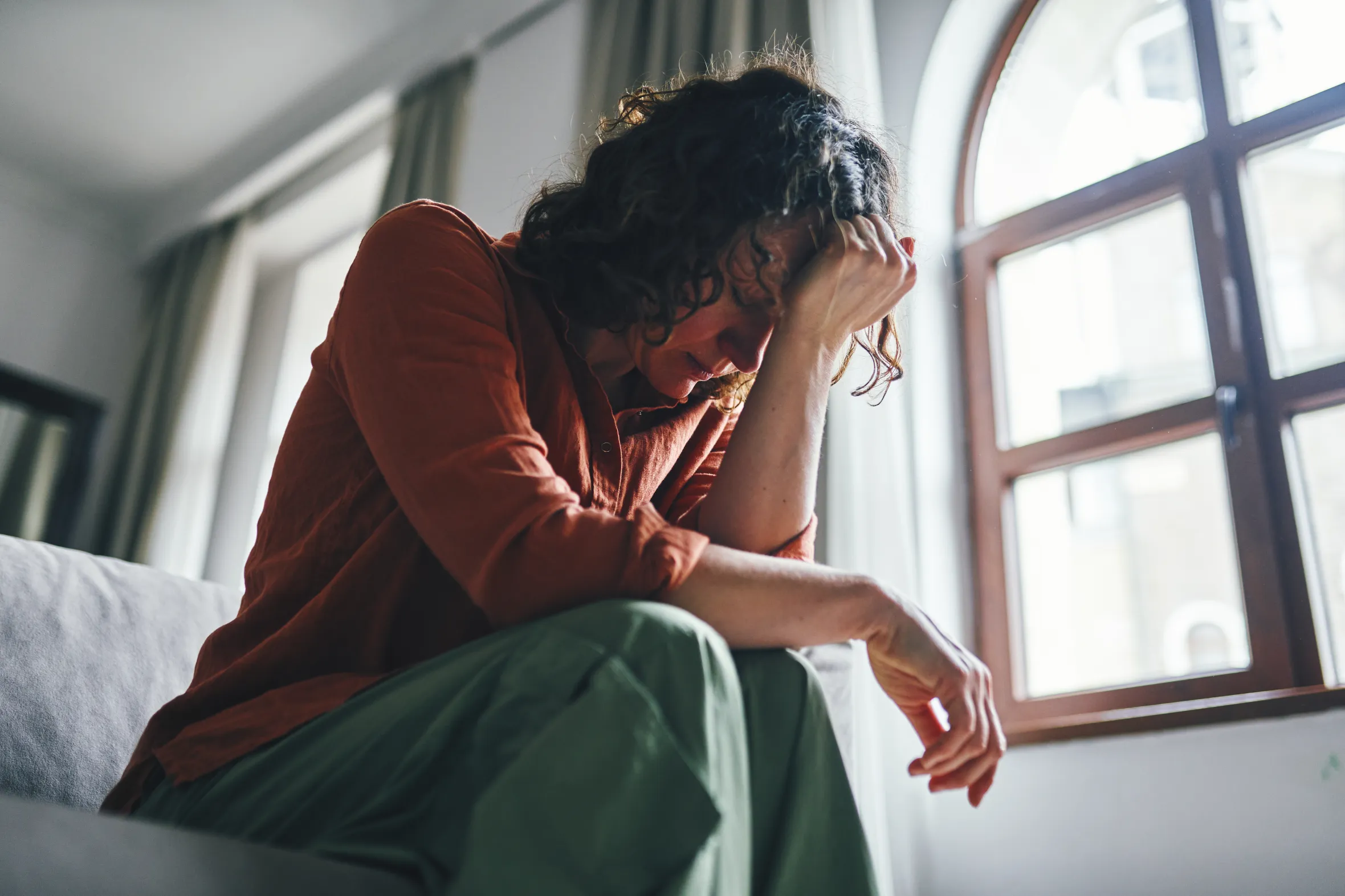 A distressed woman sitting on a sofa in a sunny apartment, leaning on her arm with an introspective expression.