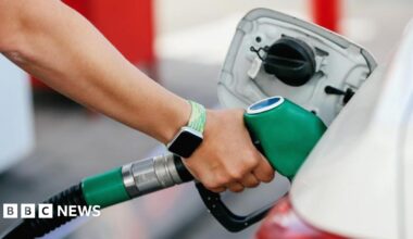 A close up stock image of an arm with a smartwatch with a light green strap putting a green unleaded petrol pump handle in to a car. The car is white.