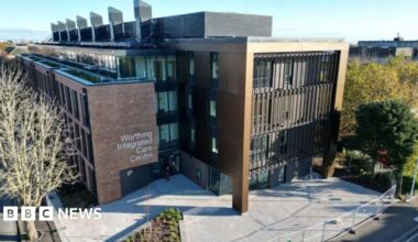 An aerial shot of the exterior of Worthing Integrated Care Centre building shows a large modern building with a sign for the centre and a paved area i front, surrounded by trees.