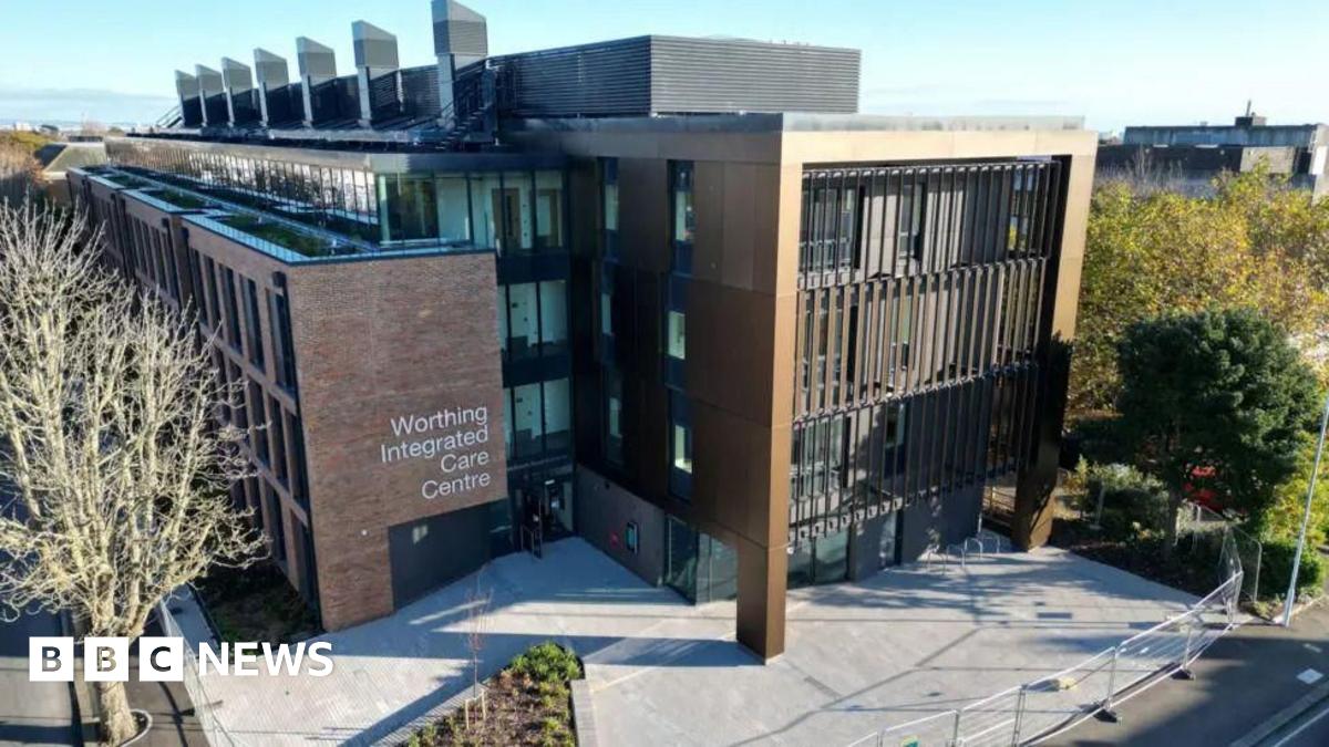 An aerial shot of the exterior of Worthing Integrated Care Centre building shows a large modern building with a sign for the centre and a paved area i front, surrounded by trees.