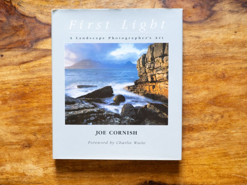 The book "First Light: A Landscape Photographer's Art" by Joe Cornish lies on a wooden surface. The cover shows a coastal landscape with rocky cliffs, waves, and distant mountains under a cloudy sky.