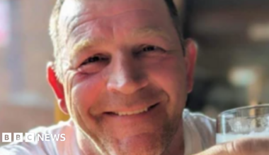 Paul Quinn, who has short light brown hair, smiles at the camera while holding a pint of what appears to be lager and wearing a white vest-top.