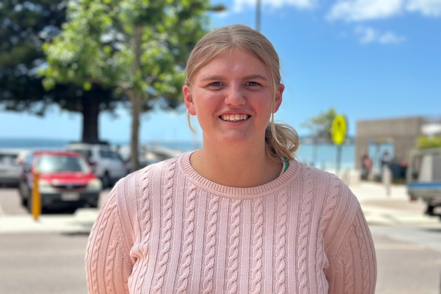 A young woman with a pink knit jumper and blonde hair tied back. Smiling at camera. Beach car park in background.