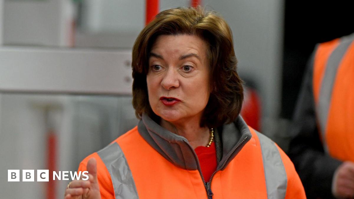 Eluned Morgan wearing a hi-viz jacket, guestering with one hand while she speaks. She is stood in front of a train which is out of focus.