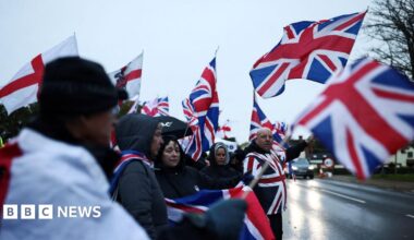 People standing next to a road opposite The Bell Hotel on an overcast day. They are waving flags showing the Union Jack and cross of St George.
