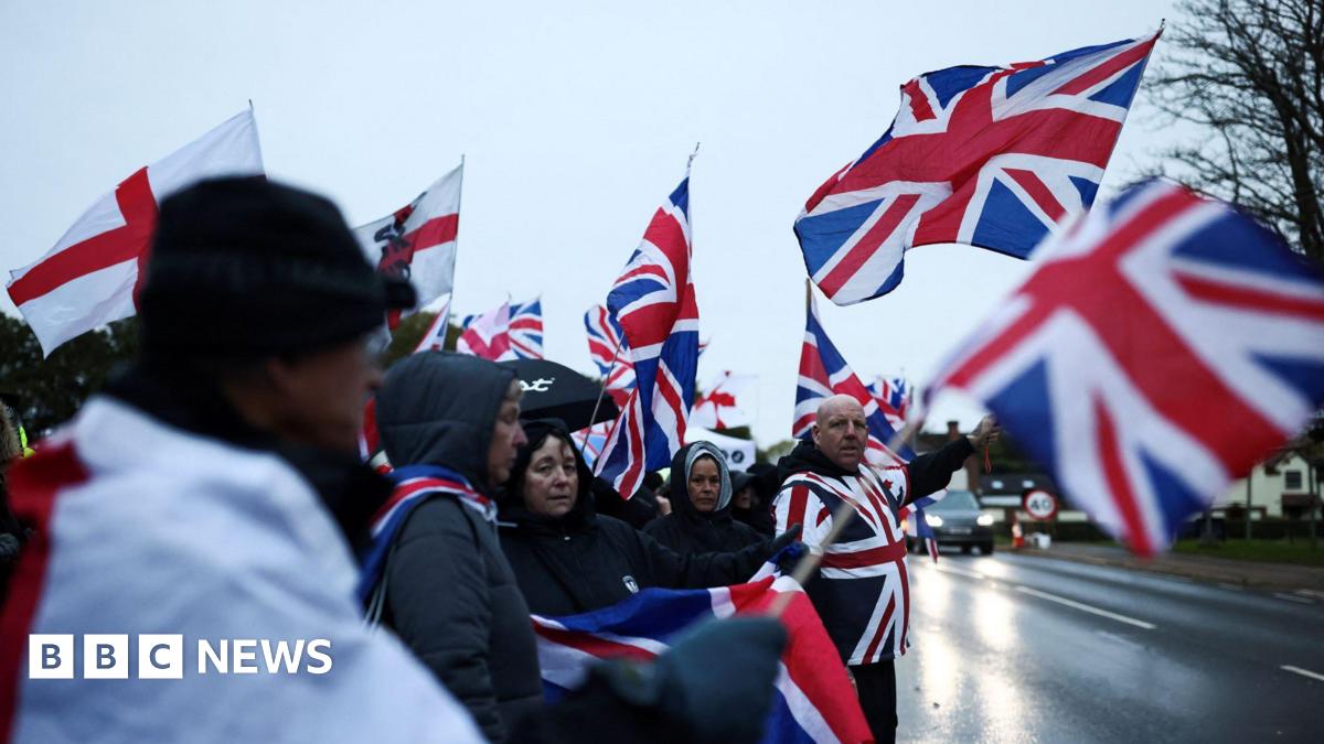 People standing next to a road opposite The Bell Hotel on an overcast day. They are waving flags showing the Union Jack and cross of St George.