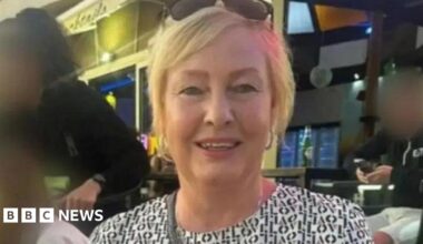 A woman in her fifties with blonde hair. She is wearing sunglasses on her head and appears to be sat in a bar. People behind her have their faces blurred.