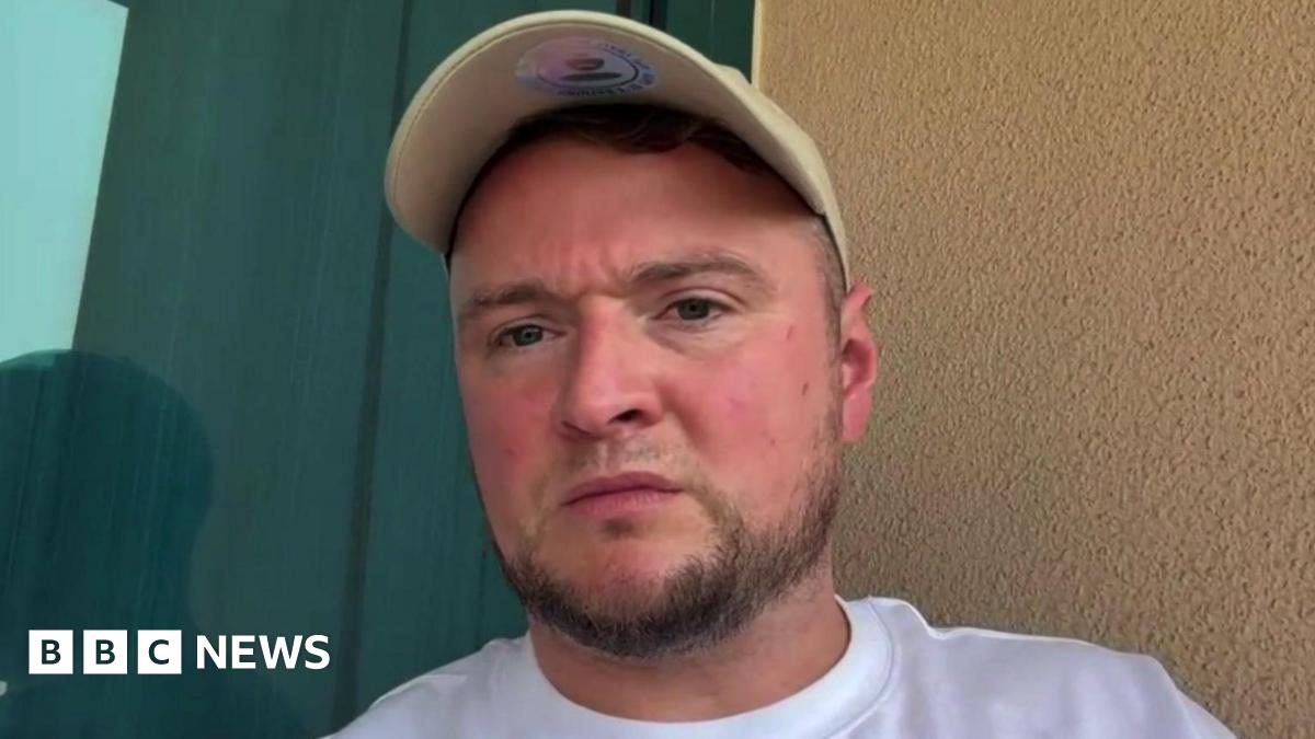A man with short brown hair sitting in a hotel room looks into a camera