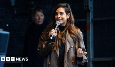 A woman in a brown leather jacket and brown hair holds a microphone