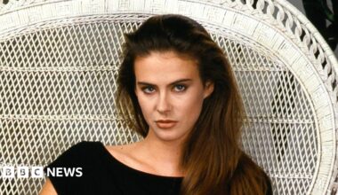 Annabel Schofield, a woman with long brown hair sits with in a white wicker chair, wearing a black dress. She looks directly at the camera with a serious expression.