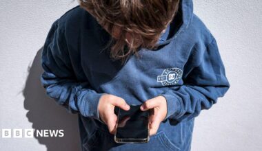 A boy in a blue hoodie leaning against a white wall, holding a phone in both hands and looking down at it. His face is covered by his brown hair.