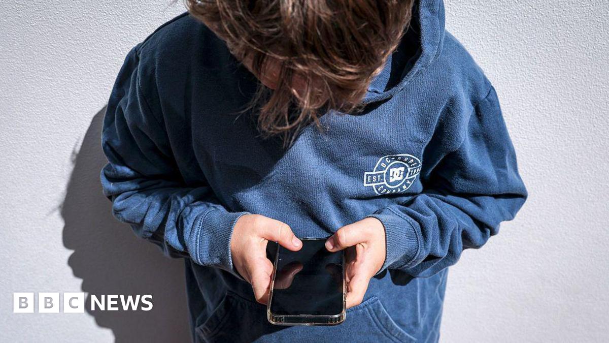 A boy in a blue hoodie leaning against a white wall, holding a phone in both hands and looking down at it. His face is covered by his brown hair.