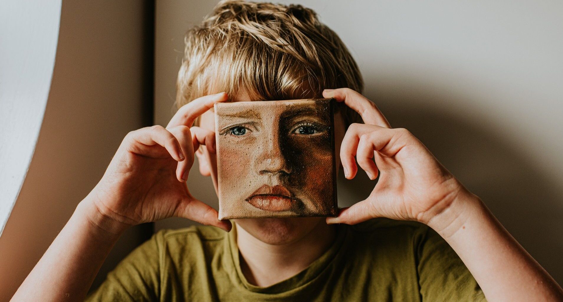 A young lad covers his face with a small oil painting of himself. The expression on the painting is serious.