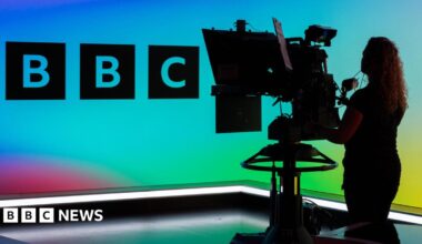 A camera operator in a TV studio, showing the BBC logo against a multi-coloured background