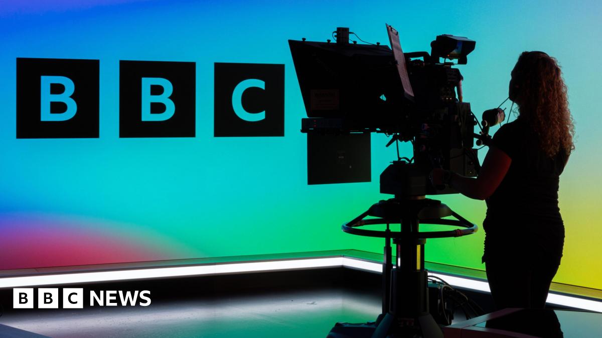 A camera operator in a TV studio, showing the BBC logo against a multi-coloured background