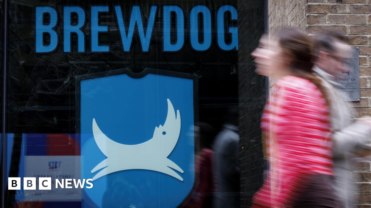 People walk past a closed-down BrewDog bar in central London. The window of the pub has the word Brewdog written in large blue letters above its logo, featuring a white dog on a blue background.
