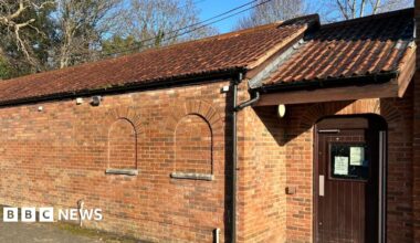 A long brick building, with a wooden door at the end. Behind there are trees peeking out above it.