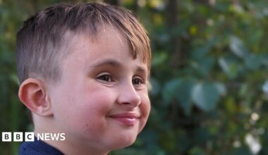 A close-up image of a young boy's face. He is looking away from the camera but smiling. There are green leaves from a tree in the background.
